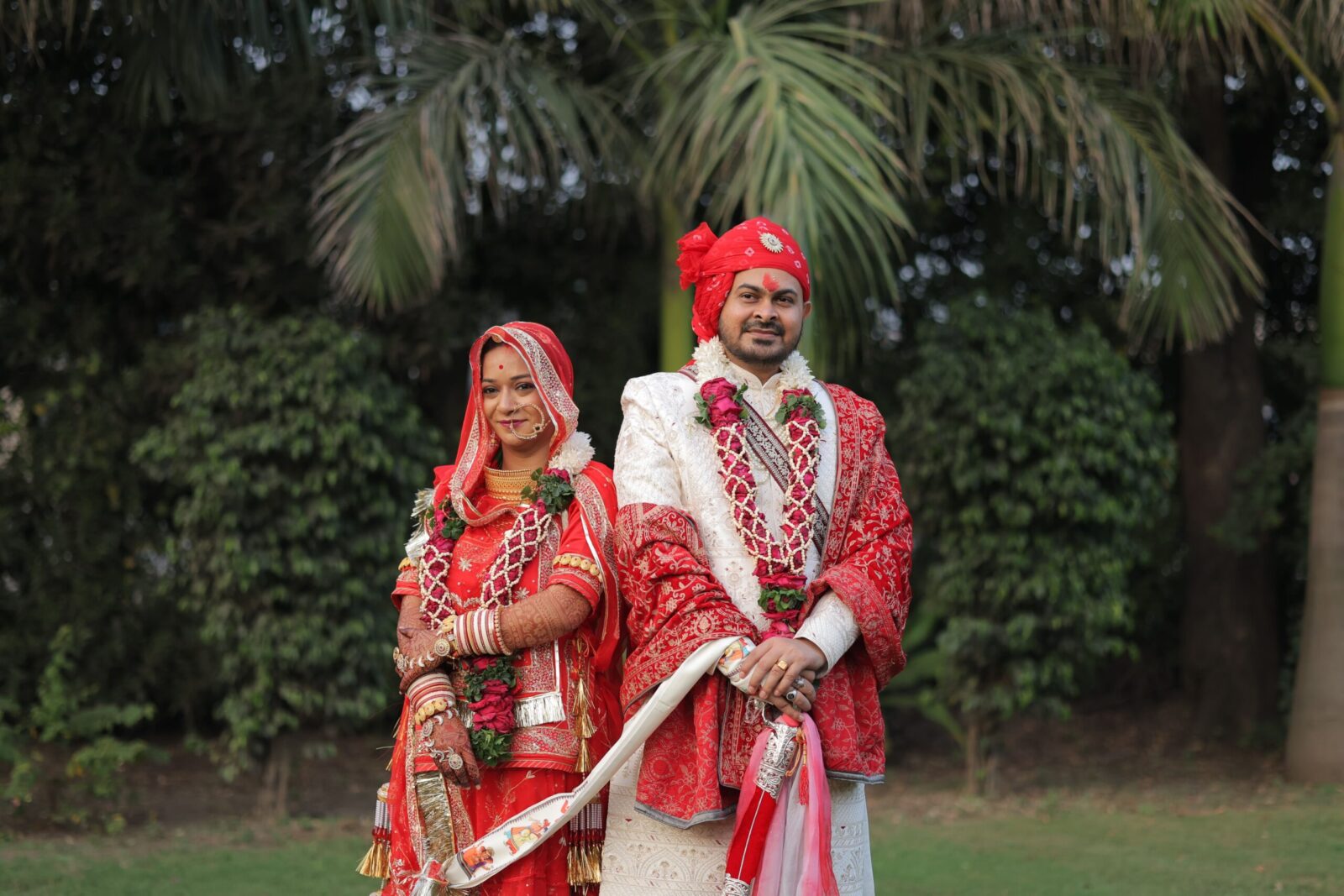 A beautiful Indian couple in traditional red attire poses for a wedding photoshoot outdoors.